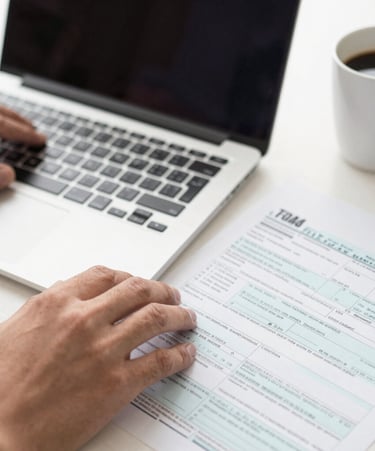 Close-up of hands typing on a laptop with tax forms and a coffee cup nearby.