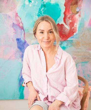 Saija Uussaari in a pink shirt and jeans sitting on a chair in front of a colorful painting