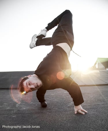 Athletic breakdancer performing a one-handed freeze on a rooftop at sunset with lens flare.