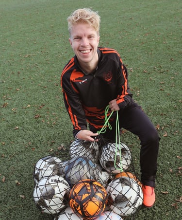 Smiling soccer coach on a turf field holding a mesh ball bag full of footballs for sports training.