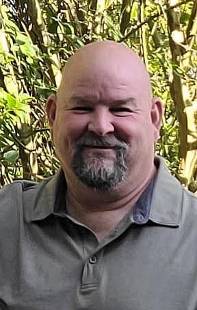 Smiling Donnie Hough with a grey goatee wearing a polo shirt outdoors against green foliage.
