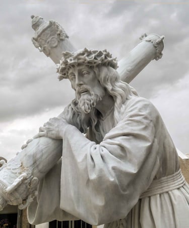 White marble statue of Jesus Christ carrying the cross with a crown of thorns against a cloudy sky.