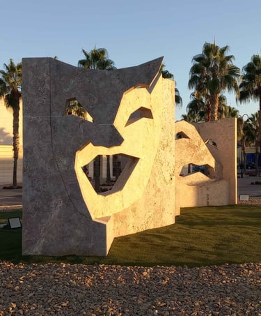 Large stone theater mask sculpture on a public roundabout with palm trees and city buildings.