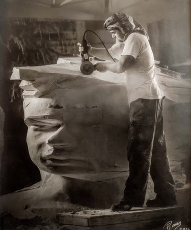 Artist using a power tool to carve a large stone head sculpture in a workshop.