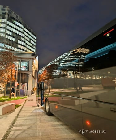 Luxury charter bus parked at night near a modern glass office building with city light reflections.