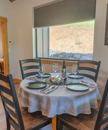 Modern rustic dining area with a round table, black chairs, and place settings near a window.