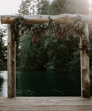Rustic wooden wedding arch with floral greenery on a lake dock surrounded by trees.