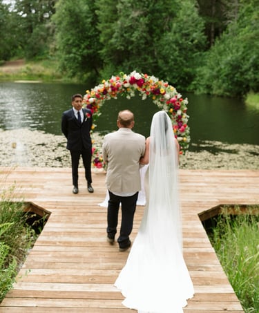 Bride in a white gown walks down a wooden pier toward a floral wedding arch by a scenic lake.