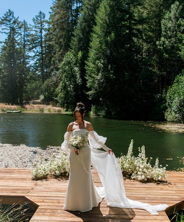 Bride in a white off-the-shoulder wedding dress posing on a wooden dock by a forest lake.