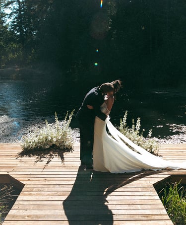 A bride and groom kiss on a wooden dock by a lake during an outdoor wedding ceremony.