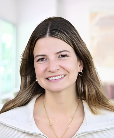 A smiling professional woman with brown hair and gold jewelry wearing a white blazer in an office.