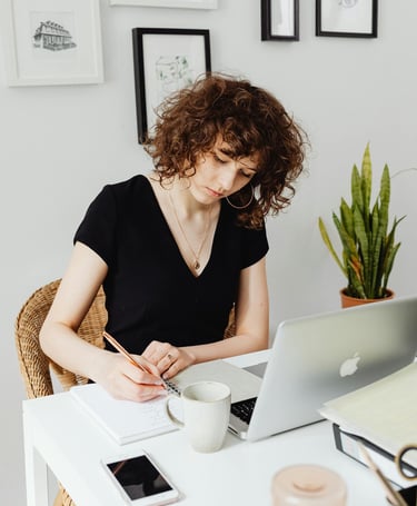 A woman in a black top sitting in front of an open laptop and wrting on a notepad