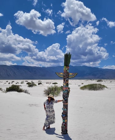 Una mujer con sombrero de sol posa junto a un colorido tótem en paisaje desértico de arena blanca.