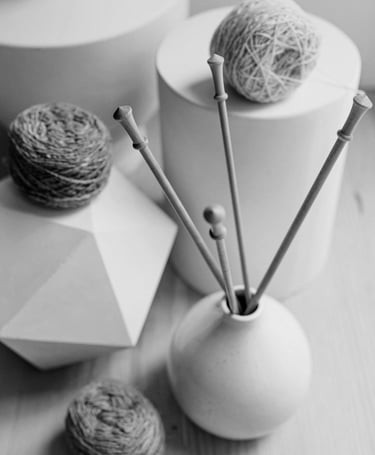Monochrome still life with textured yarn balls and knitting needles in a vase, alongside white geometric elements..