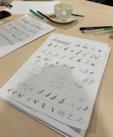 Cursive handwriting and calligraphy practice sheets on a desk with pens and a coffee cup.