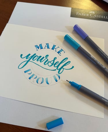 Hand-lettered blue calligraphy saying Make Yourself Proud with Faber-Castell brush markers.