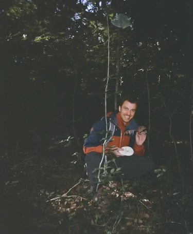Smiling hiker in a red and blue jacket crouches in a dark forest holding a mushroom