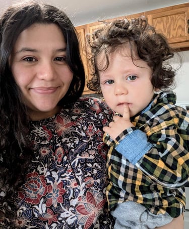 Smiling woman holding a curly-haired toddler wearing a plaid shirt in a home kitchen setting.