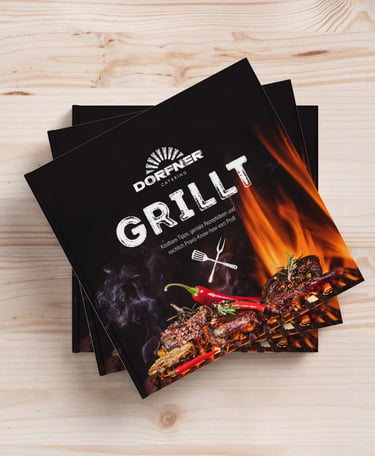 A stack of Dorfner Catering Grillt cookbooks featuring a flaming barbecue cover on a light wood background.