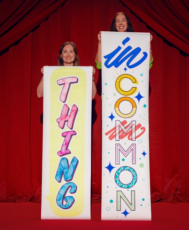 Two women holding vertical banners reading 'Thing' and 'In Common' against red theater curtains.