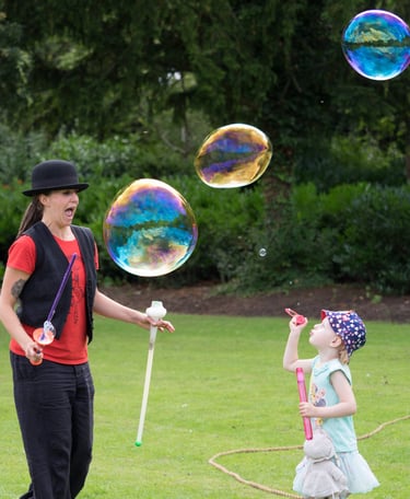 A woman and a young girl blowing large, colorful bubbles in a lush green park.
