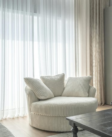 Soft linen curtains in neutral color framing a sunlit window with cozy chair beside