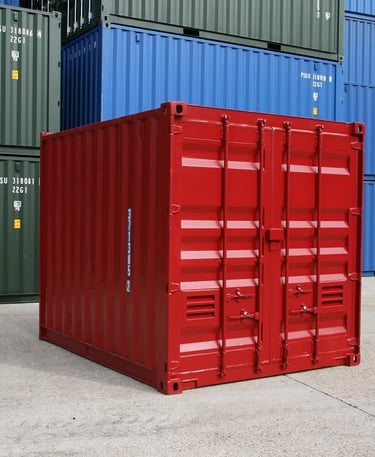 A small red metal shipping container sitting on concrete in front of blue and green cargo containers.