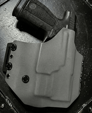 A black semi-automatic handgun secured in a custom gray Kydex OWB holster on a textured surface.