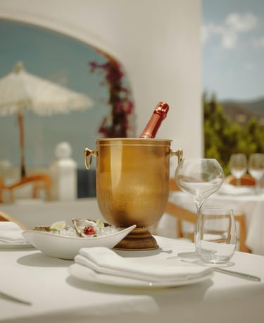Outdoor dining table with champagne in an ice bucket, oysters on a plate, and empty glasses.