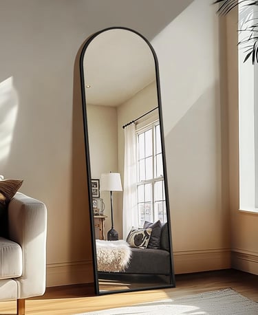 Full-length arched floor mirror with a black metal frame leaning against a wall in a bright modern living room.