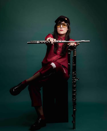A female flutist in a red outfit holding a silver flute and standing bass flute against a dark green background.