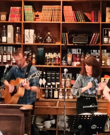 A live band performs folk music in front of a rustic bar shelf filled with vintage books and liquor bottles.