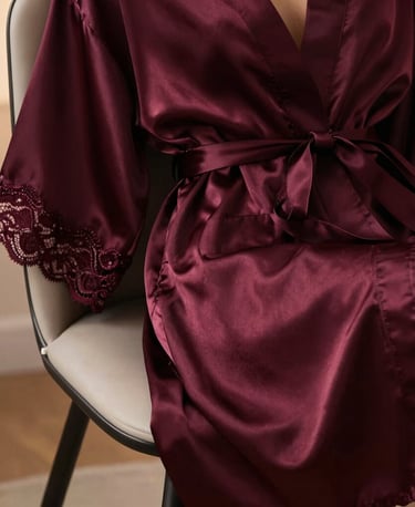 A high-fashion, intimate close-up photograph of a luxury silk robe in deep burgundy wine draped elegantly on a modern minimalist chair. Soft, romantic lighting highlights the exquisite texture of the lace trim. Warm ambient glow, sophisticated and seductive mood. Global / Western style.