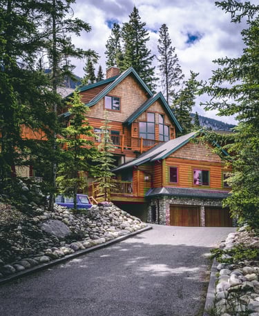 Luxury rustic log cabin with stone driveway nestled in a dense evergreen mountain forest.