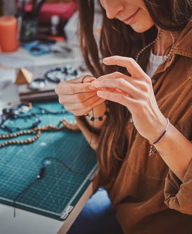 mujer sosteniendo joyeria artesanal frente a mesa de trabajo llena de cuentas e hilo