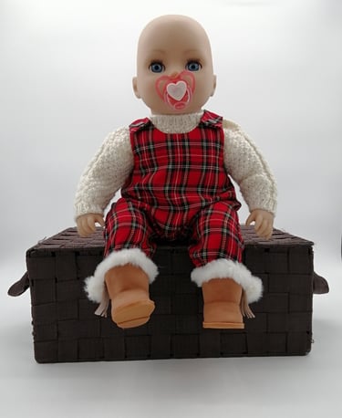 a Baby Annabell baby doll sitting on a basket dressed in handmade tartan overalls