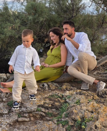 A family of three sitting on rocky terrain near a river with greenery in the background, enjoying an outdoor moment.