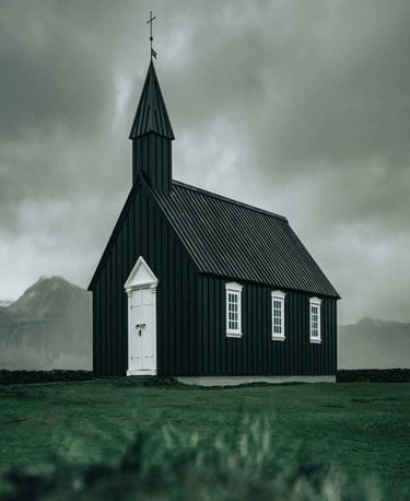 Small black church with white trim and cross-topped steeple set against dramatic cloudy sky.