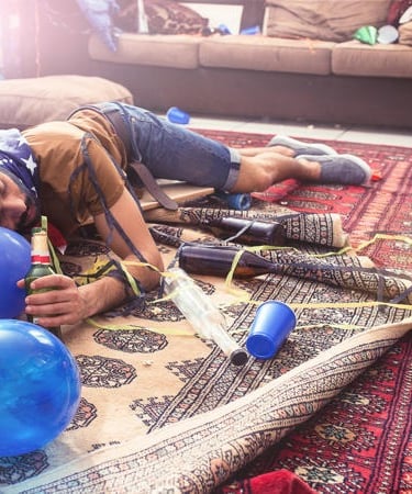 A man sleeping on a rug in a messy room after a party with balloons, beer bottles, and streamers.