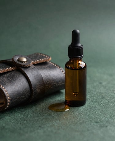 Detailed macro photograph of hand-poured organic essential oil in an amber glass dropper bottle resting beside a handcrafted leather accessory. Set in an International / Japandi Luxury sanctuary with dark forest green and earthy charcoal undertones, featuring soft shadows and minimalist natural composition.