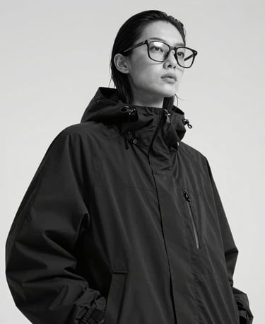 An editorial portrait of a model looking away in an oversized black technical windbreaker and structural eyewear, shot from a low angle. High contrast monochrome photography, clean studio setting, North American / International.