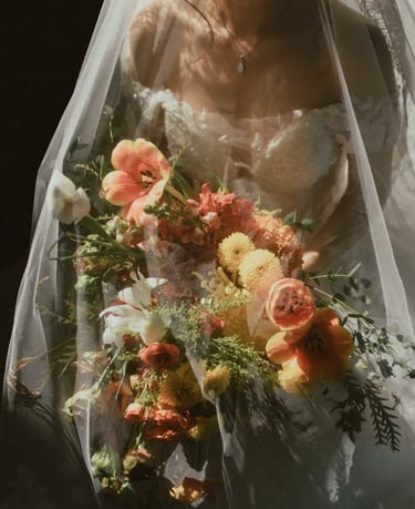 Novia con vestido de encaje bajo velo transparente sosteniendo un ramo en tonos naranja, blanco y verde.