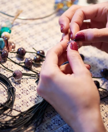 Manos de una artesana ensartando cuentas de vidrio moradas en cordón negro para crear una pulsera de macramé artesanal.
