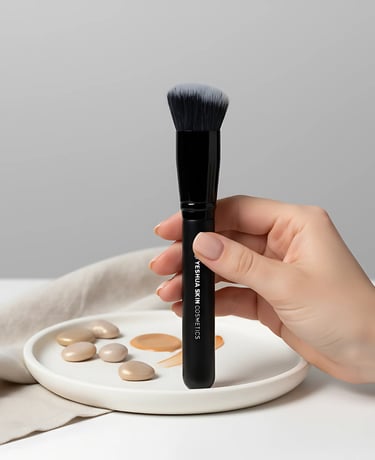 A hand holding a Yeshua Skin Cosmetics makeup foundation brush over a palette with liquid makeup drops.