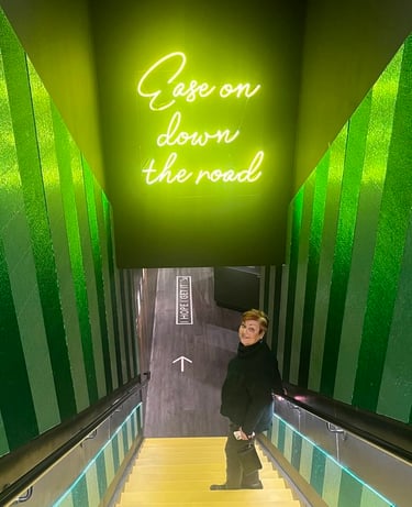 Photo of Debbie going down yellow stairs with green striped walls, standing before a neon sign: "Ease on down the road."