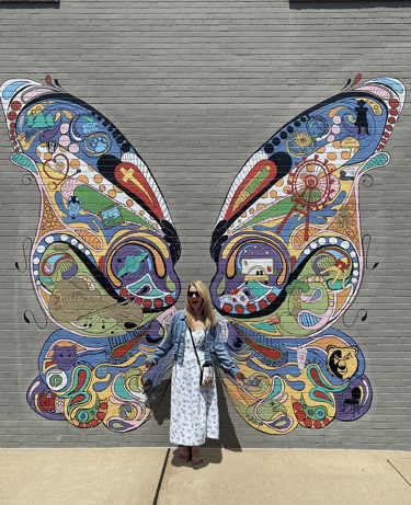 Nini standing against a wall with a mural of butterfly wings