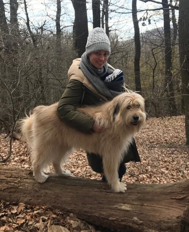 A woman in a winter hat hiking with her large fluffy dog on a fallen log in a forest.