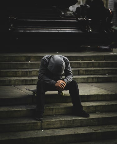 A lonely skateboarder in a grey hoodie sits on his board on city concrete stairs in a moody urban setting.
