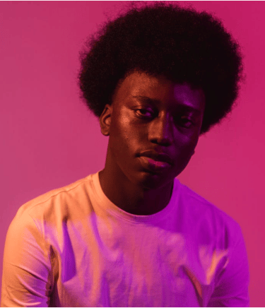 Portrait of a young Black man with an afro hairstyle under vibrant pink and purple studio lighting.
