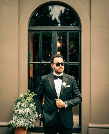 Groom in a black tuxedo and sunglasses standing confidently by a large window, by Fred Art Studio